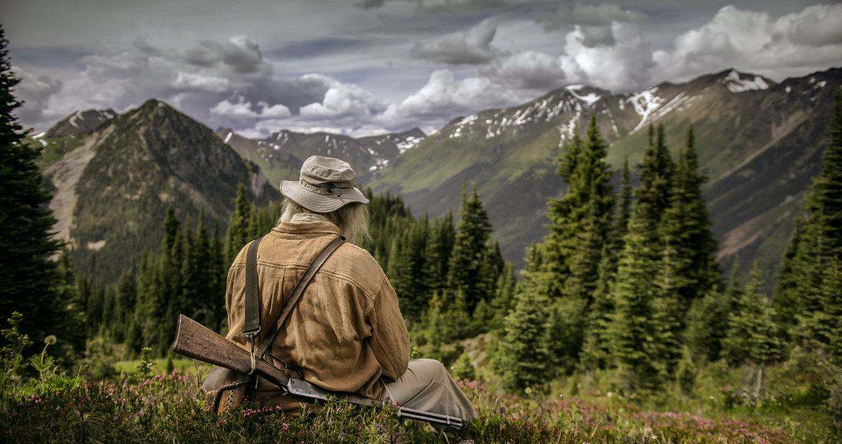 Old man sitting with his gun over his back looking at a mountain view.