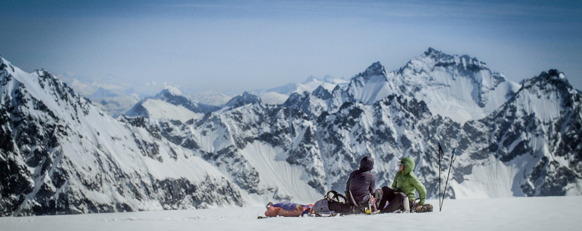 Two people sitting on snow looking out over snowy mountains